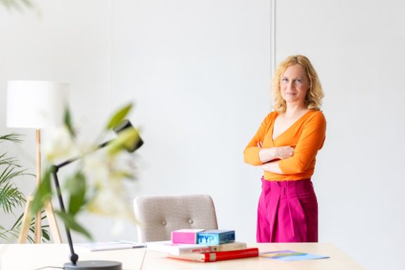 Frau mit blonden, lockigen Haaren steht in einem modernen Büro, verschränkt die Arme.